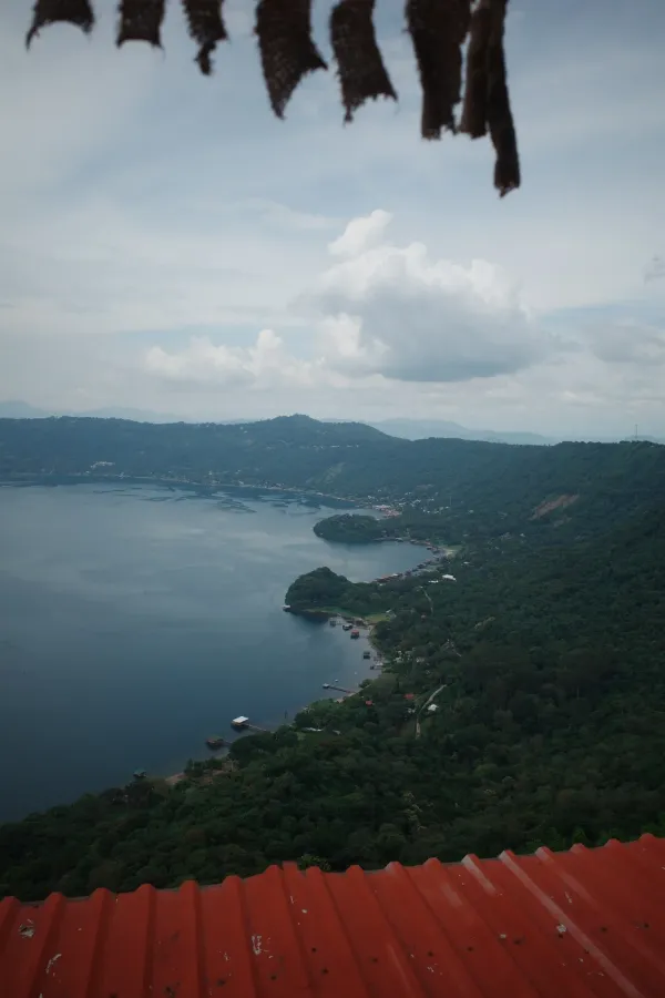 A crater lake ringed by green hills, seen from above past a red corrugated roof, with torn cloth hanging at the top of the frame.