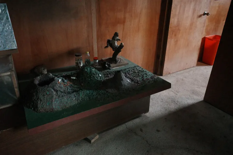 A dusty model of volcanoes on a table in a wood-paneled room, with a stuffed squirrel on a branch and a few jars behind it.