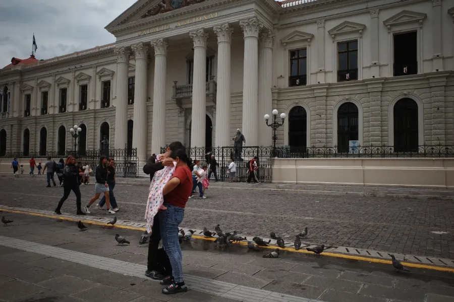 Two people embracing in a plaza in front of the Palacio Nacional, one holding a baby wrapped in a printed blanket, with pigeons at their feet.