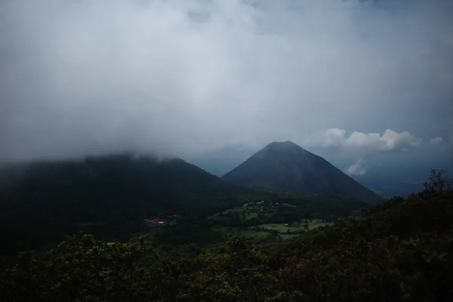 A cone-shaped volcano under heavy cloud, with a forested ridge and a small village in the valley below.