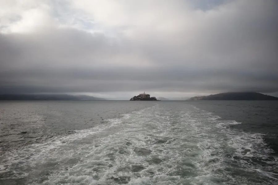 A boat's wake stretching back across grey water toward a small island under heavy cloud.
